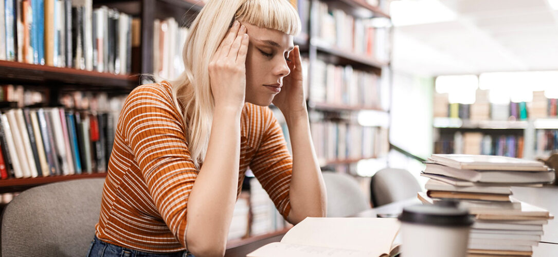 Frau sitzt gestresst in einer Bücherei oder Bibliothek über einem Buch und hält ihren Kopf mit beiden Händen an den Schläfen und scheint Kopfschmerzen zu haben.