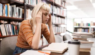 Frau sitzt gestresst in einer Bücherei oder Bibliothek über einem Buch und hält ihren Kopf mit beiden Händen an den Schläfen und scheint Kopfschmerzen zu haben.