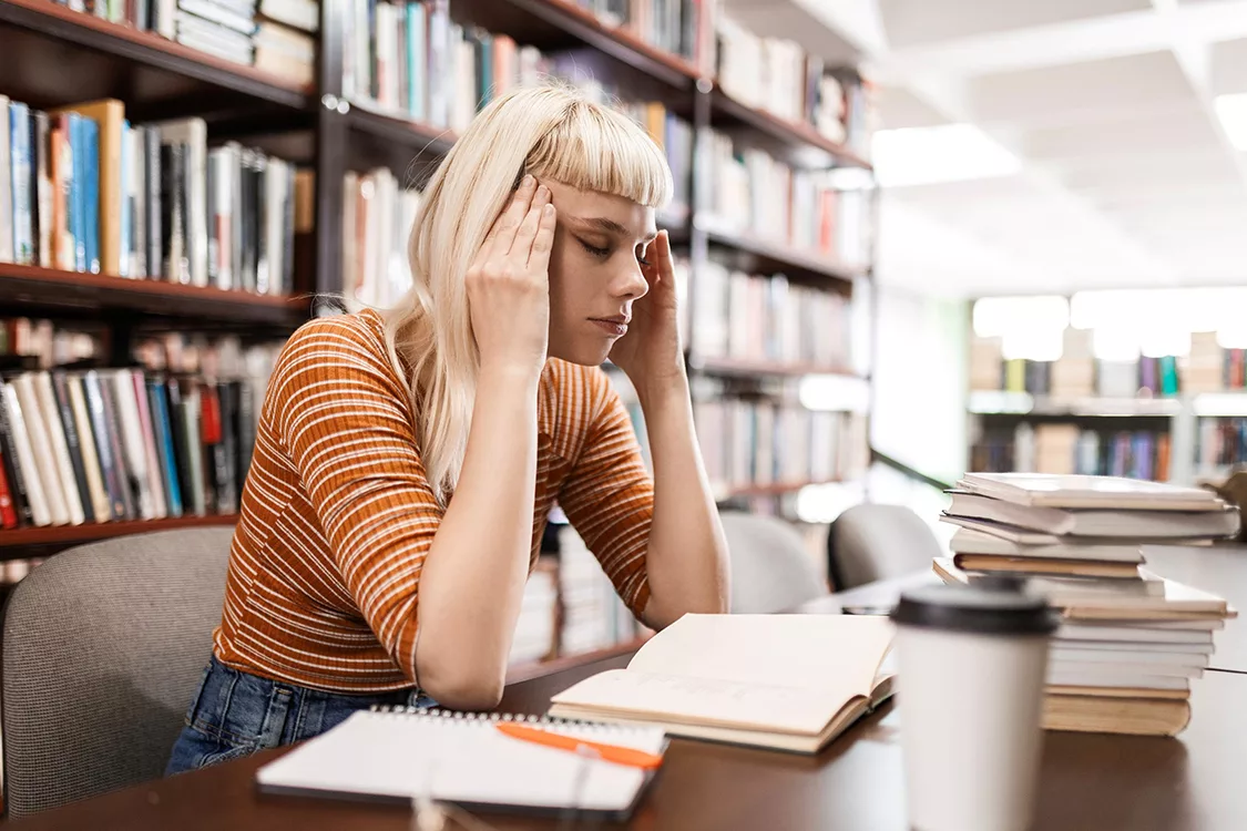 Frau sitzt gestresst in einer Bücherei oder Bibliothek über einem Buch und hält ihren Kopf mit beiden Händen an den Schläfen und scheint Kopfschmerzen zu haben.