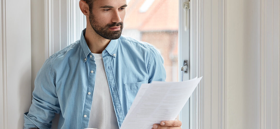 Indoor shot of serious bearded freelancer dressed in shirt, holds documentation, looks attentively at papers, does remote work, stands near window, drinks takeaway coffee. People and work concept