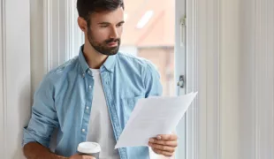 Indoor shot of serious bearded freelancer dressed in shirt, holds documentation, looks attentively at papers, does remote work, stands near window, drinks takeaway coffee. People and work concept