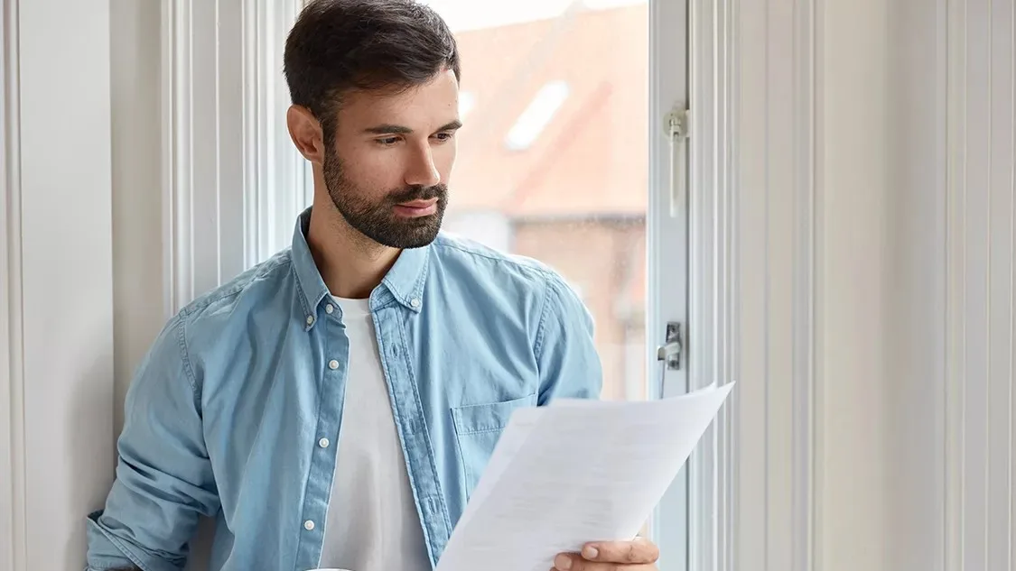 Indoor shot of serious bearded freelancer dressed in shirt, holds documentation, looks attentively at papers, does remote work, stands near window, drinks takeaway coffee. People and work concept