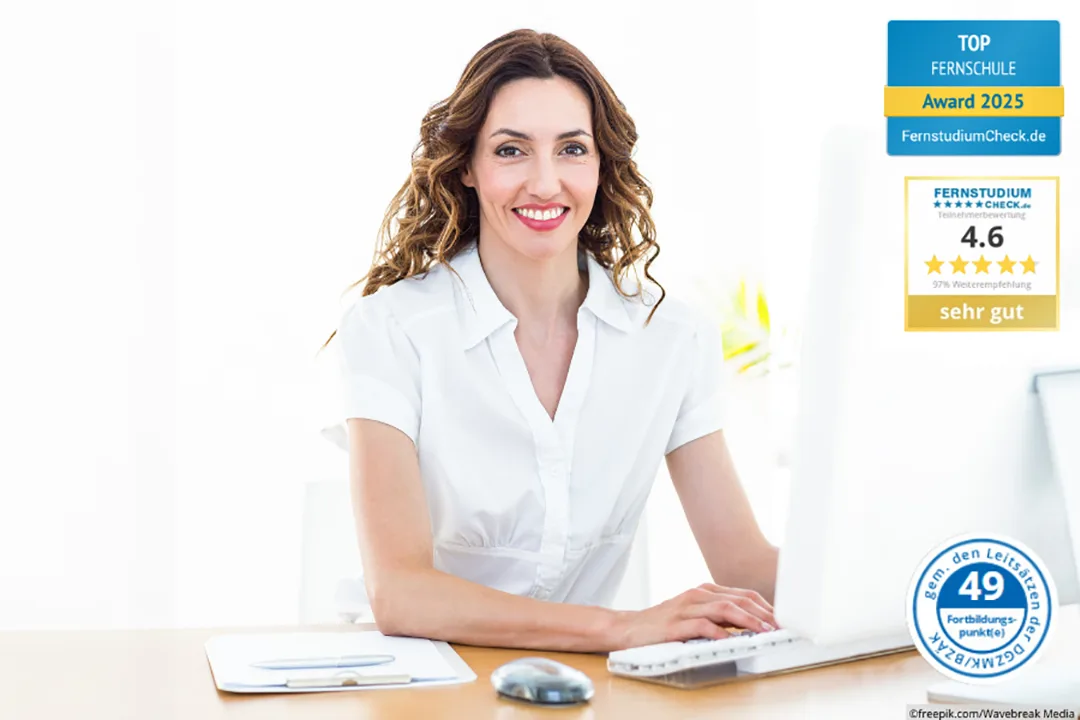 Frau mit dunklen Locken sitzt am PC und lächelt in die Kamera. Sie trägt eine weiße Bluse