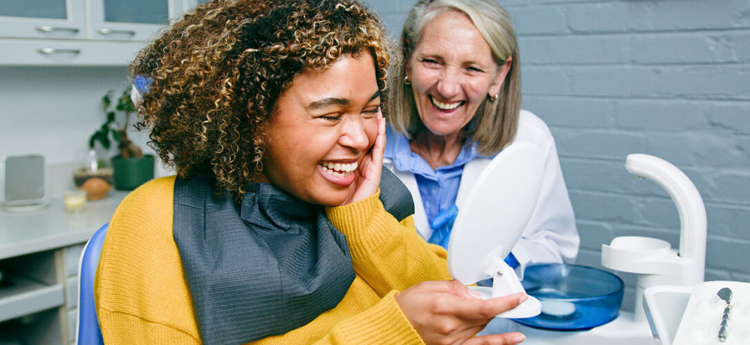 A patient and dentist share a happy moment in a dental office. The patient, seated in the dentist's chair, is looking at a handheld mirror, showcasing a joyful interaction.