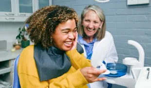 A patient and dentist share a happy moment in a dental office. The patient, seated in the dentist's chair, is looking at a handheld mirror, showcasing a joyful interaction.