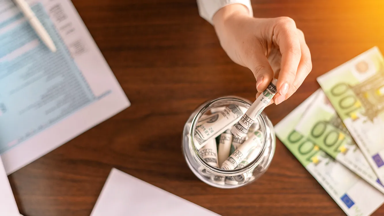 Woman dropping a banknotes into a jar with rolled banknotes on the table. Papers, money on the table