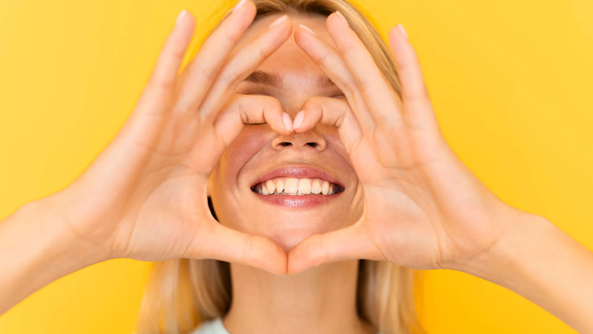 Nahaufnahme von einer Frau die lächelt und um ihren Mund herum mit ihren Händen ein Herz bildet.