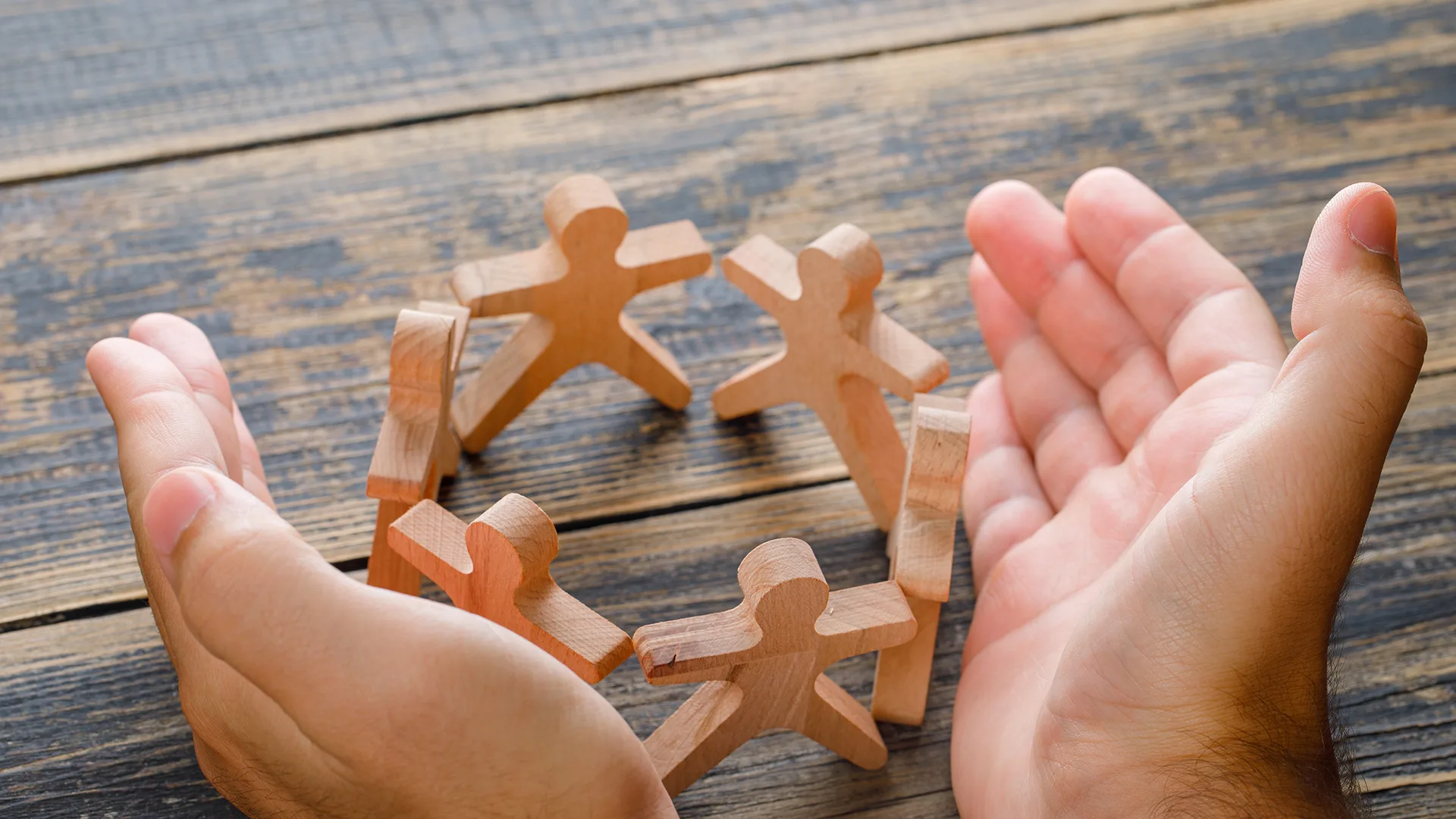 Holzfiguren bilden einen Kreis und zwei menschliche Hände umrunden sie.