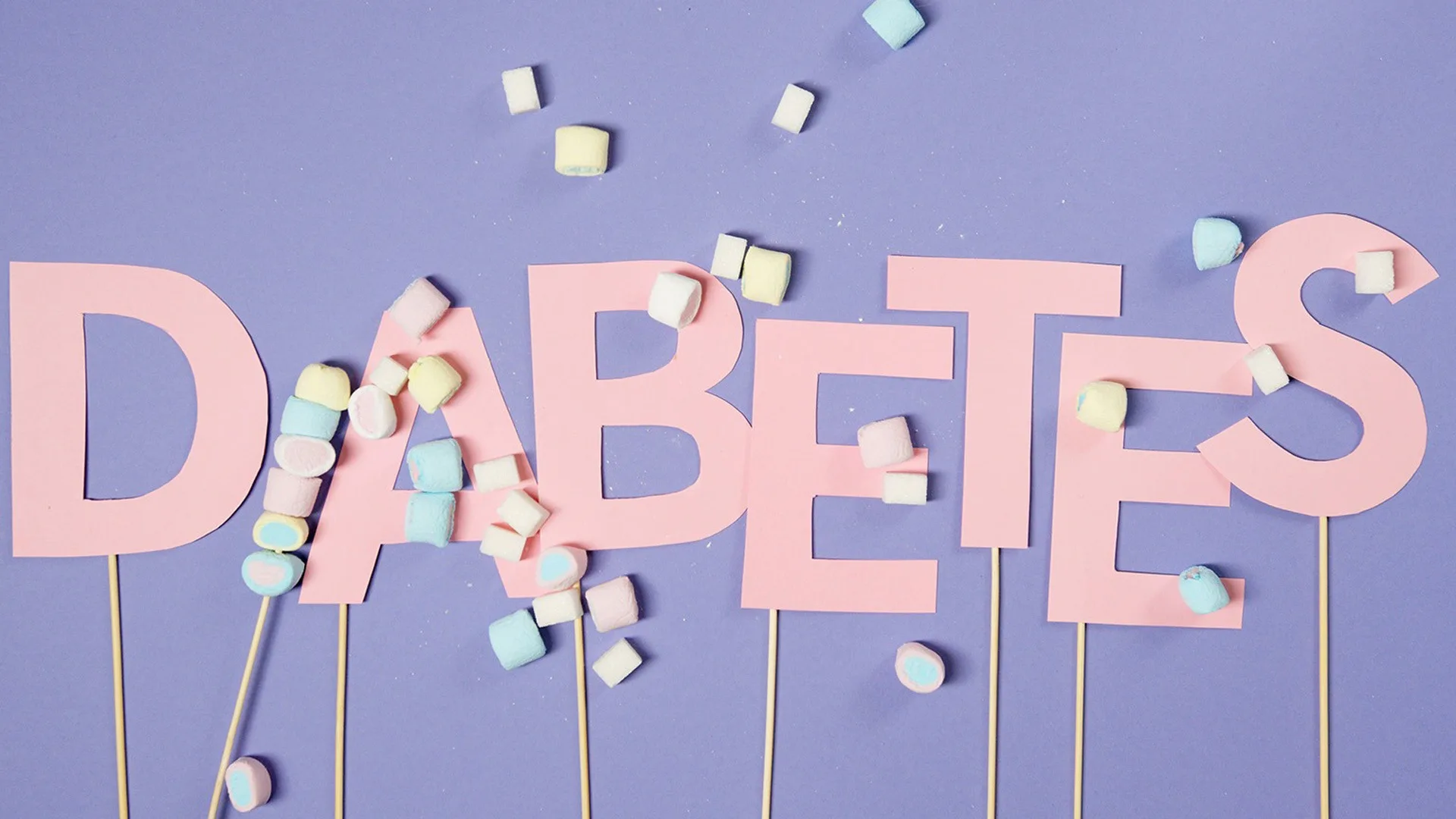 Das Wort Diabetes aus rosa Papierbuchstaben auf lila Hintergrund mit Zucker und verschiedenen Süßigkeiten umlegt.