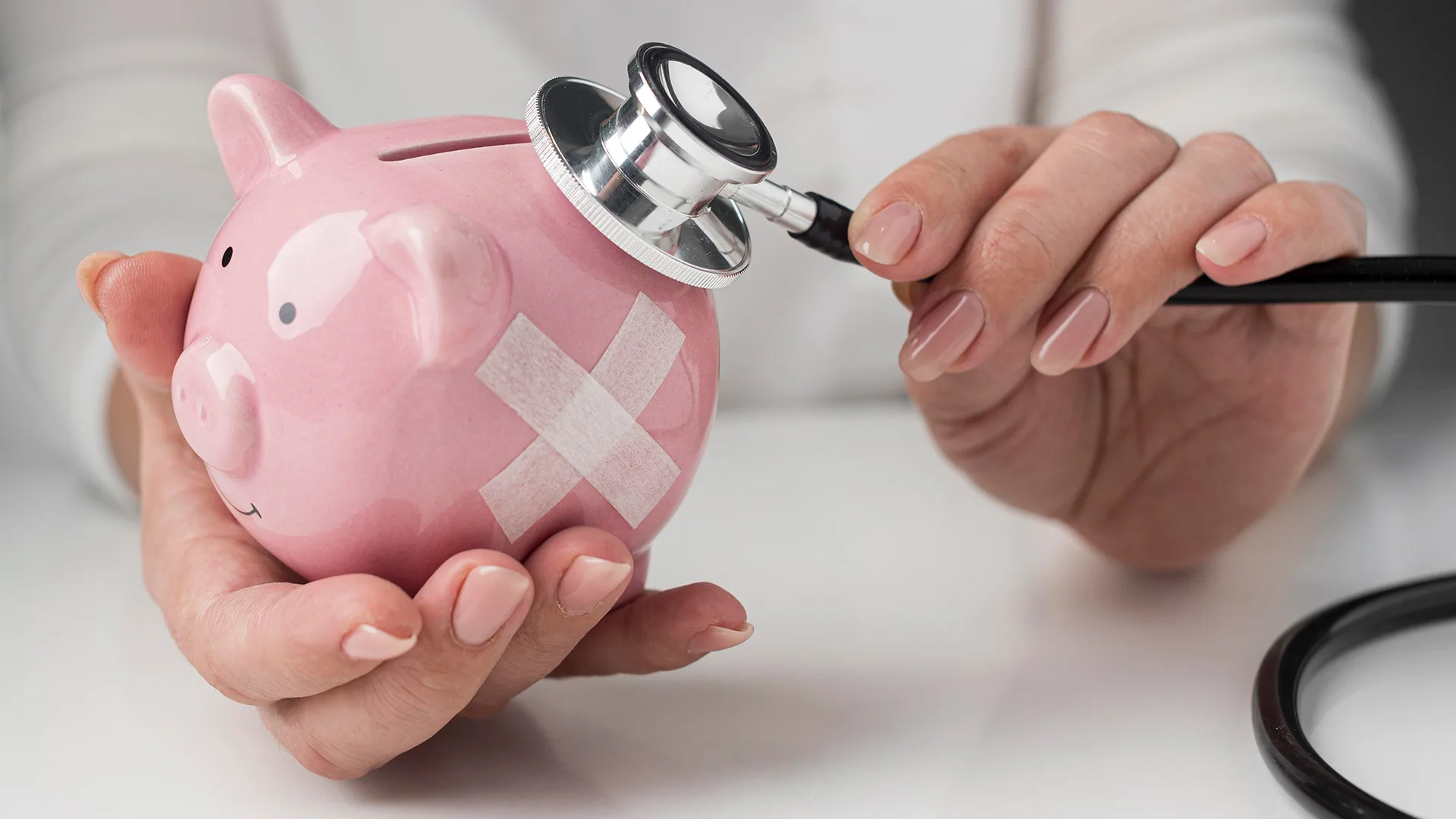 Ein Arzt hält ein Sparschwein in der Hand, mit einem Pflaster drauf und hält sein Stethoskop an das Sparschwein.