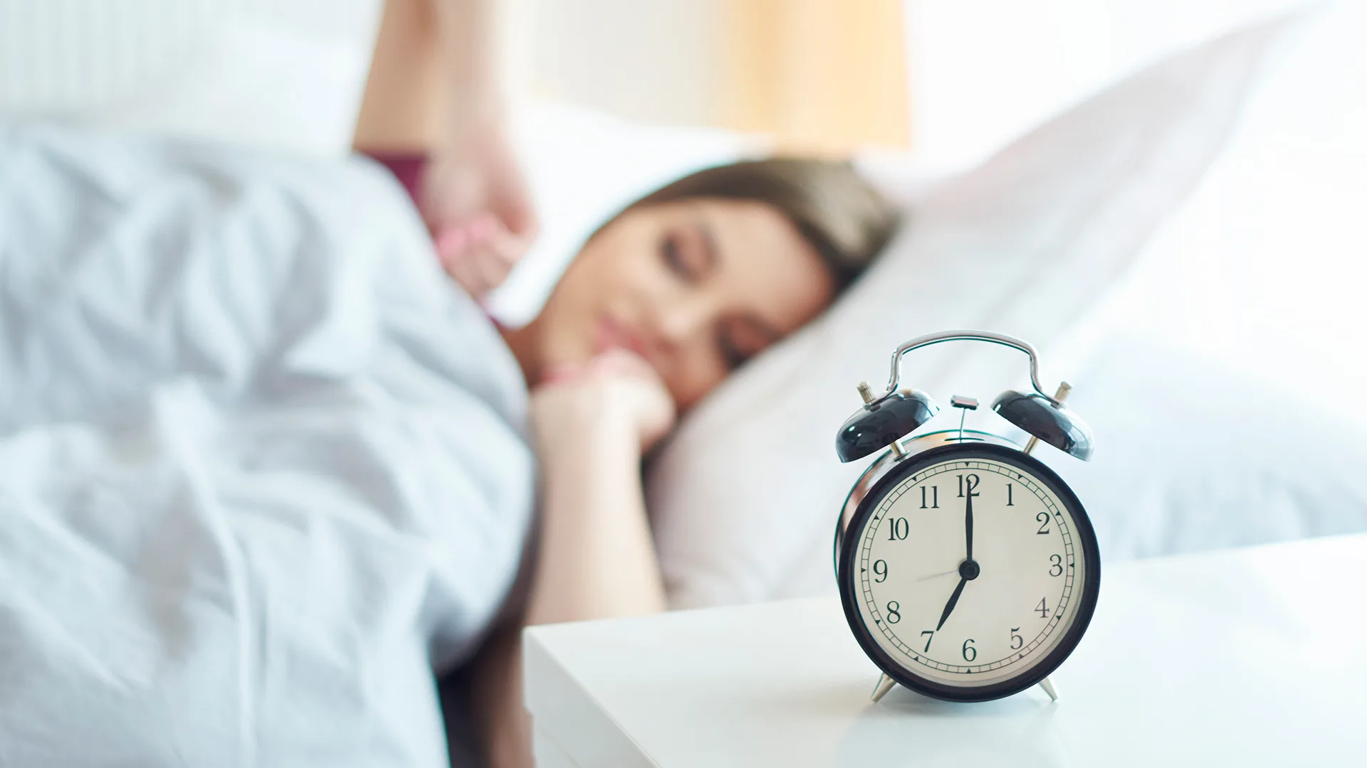 Eine Frau liegt im Bett und wacht auf, im Vordergrund eine Uhr.